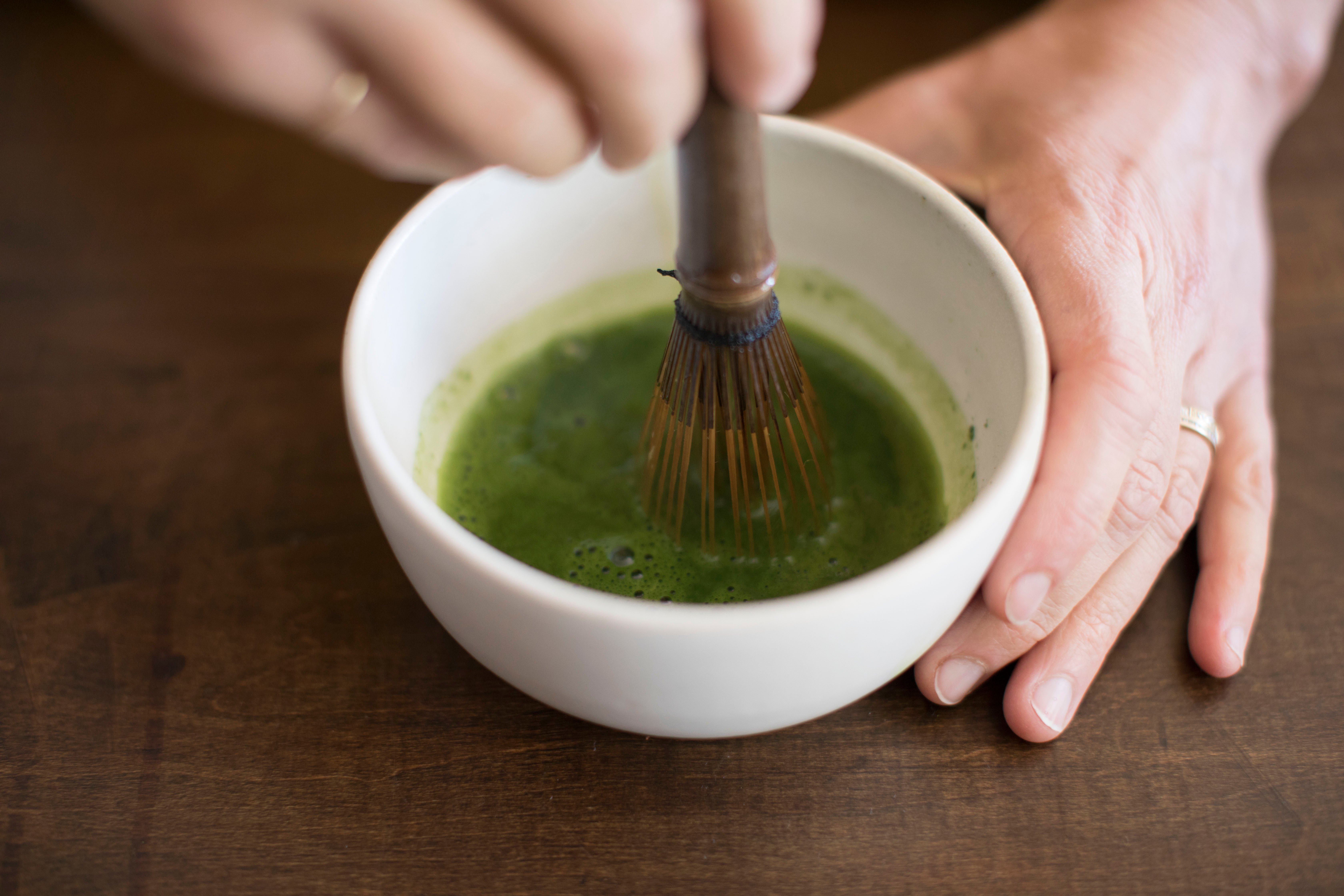 stirring green matcha in a white cup with a bamboo whisk