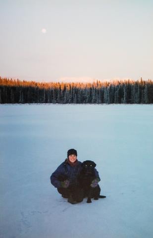 Sarah in a winter coat, hat and mittens with a black laborador retriever on a snowy lake with evergreen trees in the background